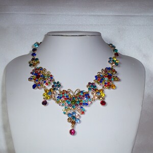 Puede incluir: Un collar de mariposa de colores con un colgante. El collar est&aacute; hecho de metal dorado y presenta una variedad de piedras de cristal de colores.