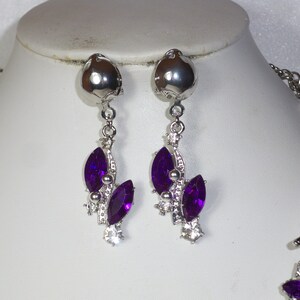 May include: A silver necklace and earrings set with purple crystal accents. The necklace has a delicate chain with a floral design and the earrings are drop style with a cluster of purple crystals.