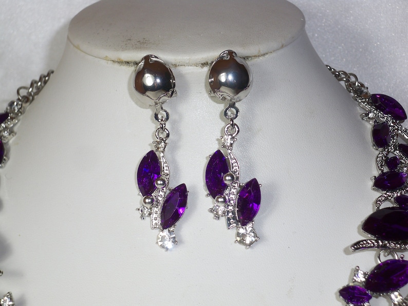 May include: A silver necklace and earrings set with purple crystal accents. The necklace has a delicate chain with a floral design and the earrings are drop style with a cluster of purple crystals.