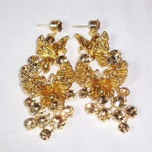 Puede incluir: Un par de pendientes de mariposa dorados. Cada pendiente presenta un dise&ntilde;o de mariposa adornado con peque&ntilde;os cristales transparentes. Racimos de cristales de color marr&oacute;n cuelgan debajo de la mariposa, a&ntilde;adiendo brillo. Los pendientes tienen cierres de poste.