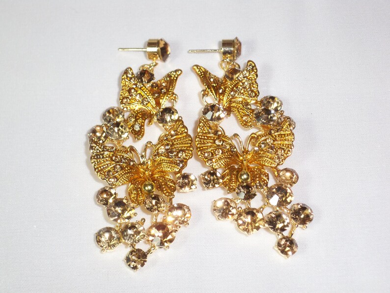 Puede incluir: Un par de pendientes de mariposa dorados. Cada pendiente presenta un dise&ntilde;o de mariposa adornado con peque&ntilde;os cristales transparentes. Racimos de cristales de color marr&oacute;n cuelgan debajo de la mariposa, a&ntilde;adiendo brillo. Los pendientes tienen cierres de poste.