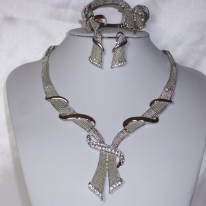 May include: A silver-toned jewelry set displayed on a mannequin. The set includes a necklace, bracelet, and earrings. The necklace and bracelet feature a mesh design with curved accents and are embellished with clear crystals. The earrings are a matching design.