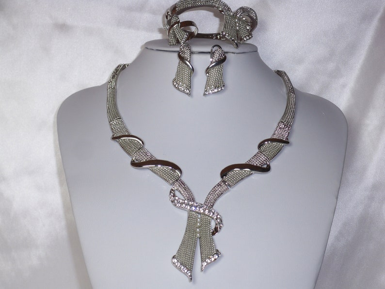 May include: A silver-toned jewelry set displayed on a mannequin. The set includes a necklace, bracelet, and earrings. The necklace and bracelet feature a mesh design with curved accents and are embellished with clear crystals. The earrings are a matching design.