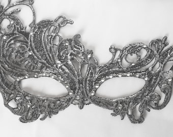 May include: A black lace masquerade mask featuring intricate swirling embroidery patterns and small, clear, reflective rhinestones embedded along the eye openings and across the detailed filigree lace structure of the accessory.