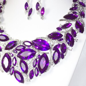 May include: A silver necklace with a purple crystal design. The necklace features a large, ornate pendant with multiple purple crystals arranged in a floral pattern. The necklace is accented with smaller, clear crystals.