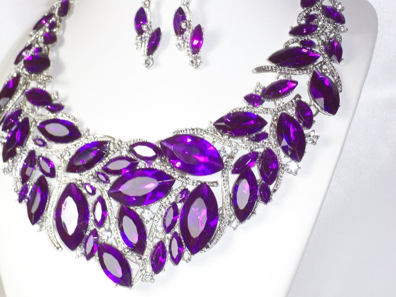 May include: A silver necklace with a purple crystal design. The necklace features a large, ornate pendant with multiple purple crystals arranged in a floral pattern. The necklace is accented with smaller, clear crystals.