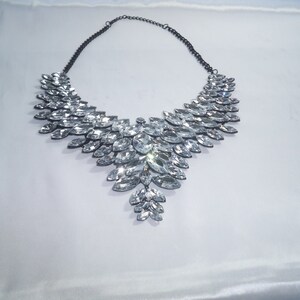 Puede incluir: Un collar llamativo con una cadena de metal oscuro y un gran colgante en forma de V, compuesto por numerosos cristales transparentes de talla marquesa dispuestos en un patr&oacute;n de hojas superpuestas que se estrecha hacia el centro inferior.