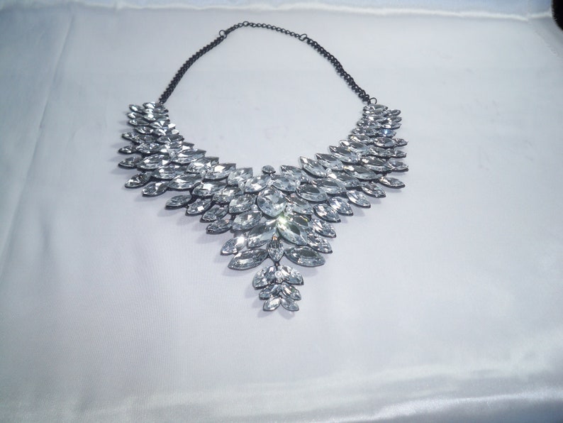 Puede incluir: Un collar llamativo con una cadena de metal oscuro y un gran colgante en forma de V, compuesto por numerosos cristales transparentes de talla marquesa dispuestos en un patr&oacute;n de hojas superpuestas que se estrecha hacia el centro inferior.