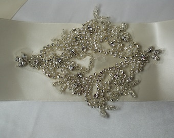 May include: A white satin ribbon with a large, ornate embellishment made of silver beads and rhinestones. The embellishment is in the shape of a diamond and has a delicate, intricate design.