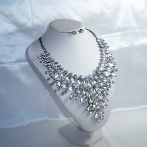 Puede incluir: Un collar llamativo con numerosos cristales transparentes de talla marquesa dispuestos en un patr&oacute;n de hojas superpuestas sobre una cadena de metal oscuro, acompa&ntilde;ado de un par de pendientes de bot&oacute;n de cristal a juego sobre un busto blanco.