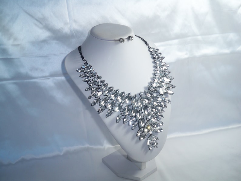 Puede incluir: Un collar llamativo con numerosos cristales transparentes de talla marquesa dispuestos en un patr&oacute;n de hojas superpuestas sobre una cadena de metal oscuro, acompa&ntilde;ado de un par de pendientes de bot&oacute;n de cristal a juego sobre un busto blanco.
