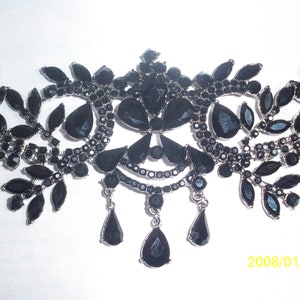 May include: An ornate black necklace with a silver-tone metal frame. The necklace features a symmetrical design with leaf-shaped and teardrop-shaped black gemstones. The design includes dangling teardrop pendants.