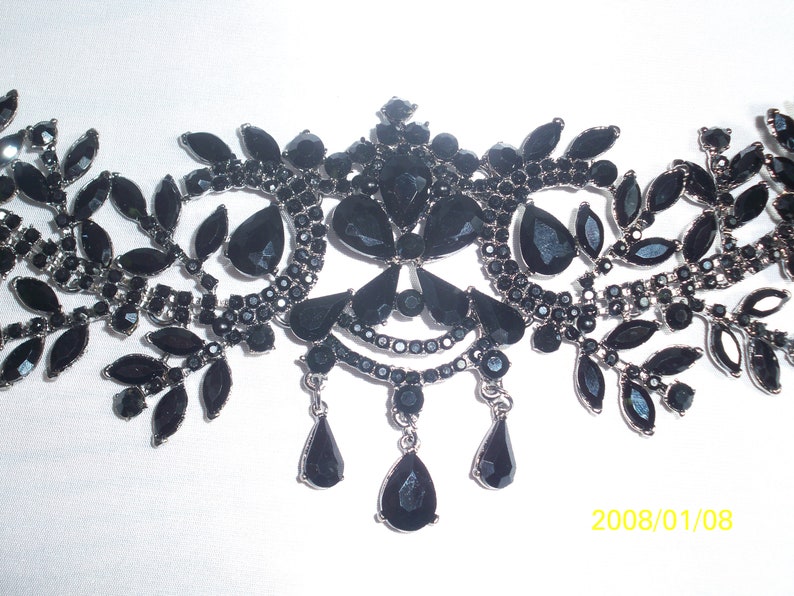 May include: An ornate black necklace with a silver-tone metal frame. The necklace features a symmetrical design with leaf-shaped and teardrop-shaped black gemstones. The design includes dangling teardrop pendants.