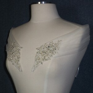 May include: A white off-the-shoulder wedding dress with a sheer overlay and beaded embellishments on the shoulders.