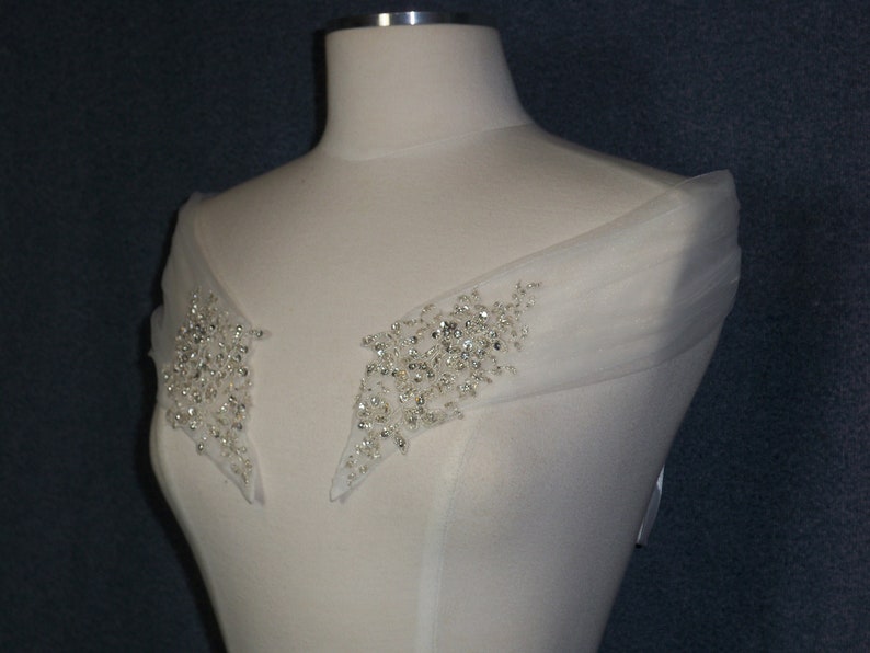May include: A white off-the-shoulder wedding dress with a sheer overlay and beaded embellishments on the shoulders.