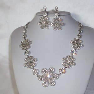May include: A silver-toned necklace and earring set featuring multiple flower-shaped charms encrusted with clear rhinestones. The necklace consists of a chain of these floral links, while the matching earrings feature a single flower drop design.