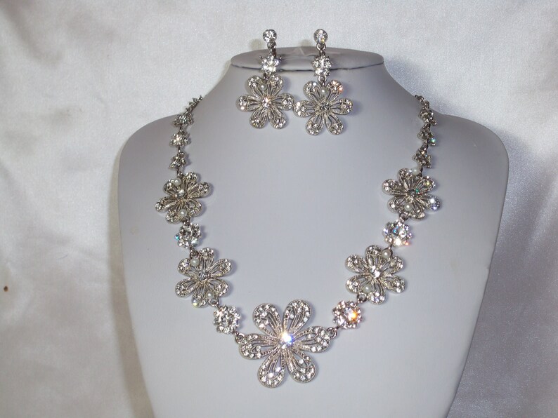 May include: A silver-toned necklace and earring set featuring multiple flower-shaped charms encrusted with clear rhinestones. The necklace consists of a chain of these floral links, while the matching earrings feature a single flower drop design.