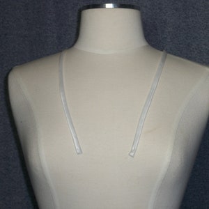 May include: A cream-colored dress form with a silver-colored metal neck and two thin, white straps. The dress form is set against a textured, dark gray background.