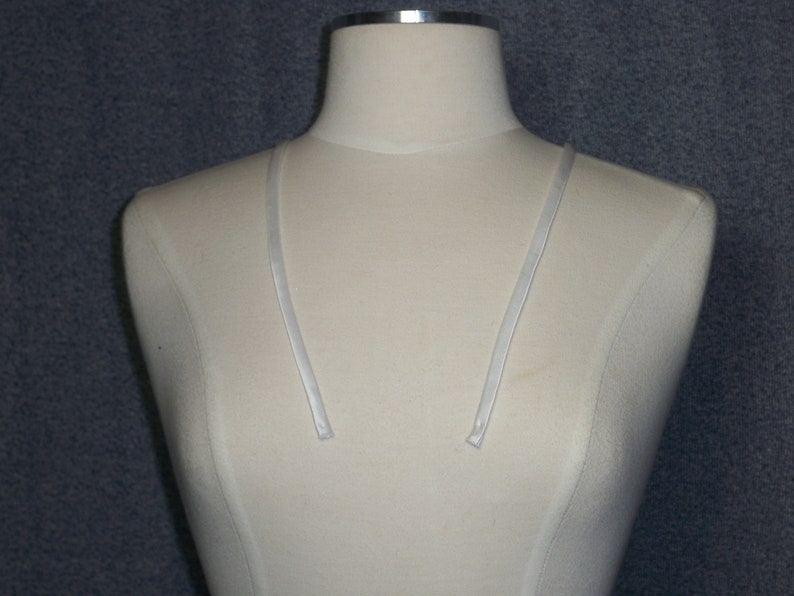 May include: A cream-colored dress form with a silver-colored metal neck and two thin, white straps. The dress form is set against a textured, dark gray background.
