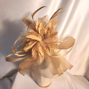 以下が含まれることがあります： ベージュ色のファシネーターヘッドピース。中央に布製の花びら、質感のあるメッシュネット、繊細な羽があしらわれています。小さな真珠のようなアクセントと、土台部分には透け感のある布で作られた幅広で立体的なループが施されています。