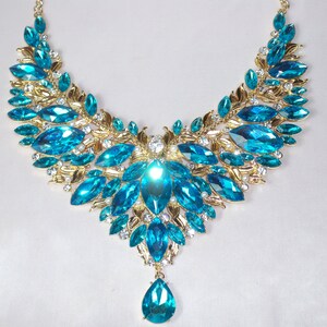 May include: A statement necklace featuring large, teardrop-shaped, and marquise-cut turquoise-colored gemstones. The gemstones are set in a gold-toned metal with leaf and floral designs, and small clear crystals.