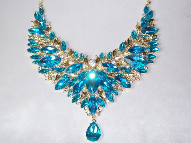 May include: A statement necklace featuring large, teardrop-shaped, and marquise-cut turquoise-colored gemstones. The gemstones are set in a gold-toned metal with leaf and floral designs, and small clear crystals.