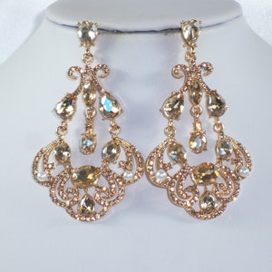May include: A pair of gold-toned chandelier earrings. Each earring features multiple teardrop and oval-shaped crystals in shades of champagne and clear, with small pearl accents. The earrings have an ornate, filigree design.