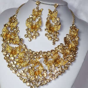 Puede incluir: Un conjunto de collar y pendientes dorados. El collar y los pendientes presentan dise&ntilde;os de mariposas y est&aacute;n adornados con cristales transparentes y brillantes. Las joyas se exhiben sobre un busto de maniqu&iacute; blanco sobre un fondo blanco.