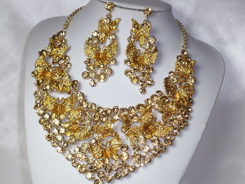 Puede incluir: Un conjunto de collar y pendientes dorados. El collar y los pendientes presentan dise&ntilde;os de mariposas y est&aacute;n adornados con cristales transparentes y brillantes. Las joyas se exhiben sobre un busto de maniqu&iacute; blanco sobre un fondo blanco.