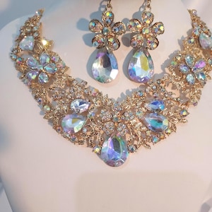 May include: A gold-coloured jewellery set comprising a necklace and matching earrings. The necklace and earrings feature iridescent, teardrop-shaped gemstones and floral details. The set is displayed on a white mannequin bust.