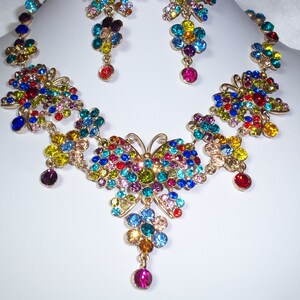 Puede incluir: Un collar de oro con un dise&ntilde;o de mariposa presenta piedras de imitaci&oacute;n de colores en tonos de rojo, azul, verde, amarillo y morado. El collar tiene un colgante con un dise&ntilde;o de mariposa m&aacute;s grande.