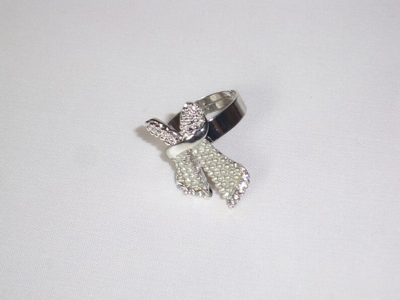 May include: Silver-toned ring featuring a bow design. The bow is adorned with small, clear rhinestones and faux pearls, with a white enamel accent. The ring is set against a white background.