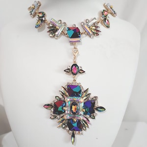 May include: A dazzling statement necklace featuring a gold-toned chain adorned with iridescent, multi-faceted crystals. The pendant showcases a large, ornate cross design with a central, rectangular crystal and surrounding smaller gems. The necklace is displayed on a white mannequin.