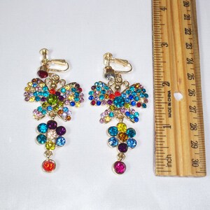 Puede incluir: Un par de pendientes de clip con un dise&ntilde;o de mariposa adornado con brillantes de colores. Las mariposas est&aacute;n adornadas con colgantes con forma de flor, tambi&eacute;n adornados con brillantes.
