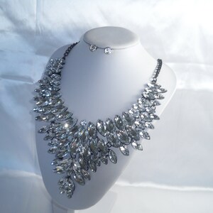 Puede incluir: Un collar llamativo con numerosos cristales transparentes de talla marquesa dispuestos en un patr&oacute;n denso y superpuesto. Los cristales est&aacute;n engastados en un marco de metal oscuro con una cadena a juego. Se incluye un par de pendientes de cristal redondos.