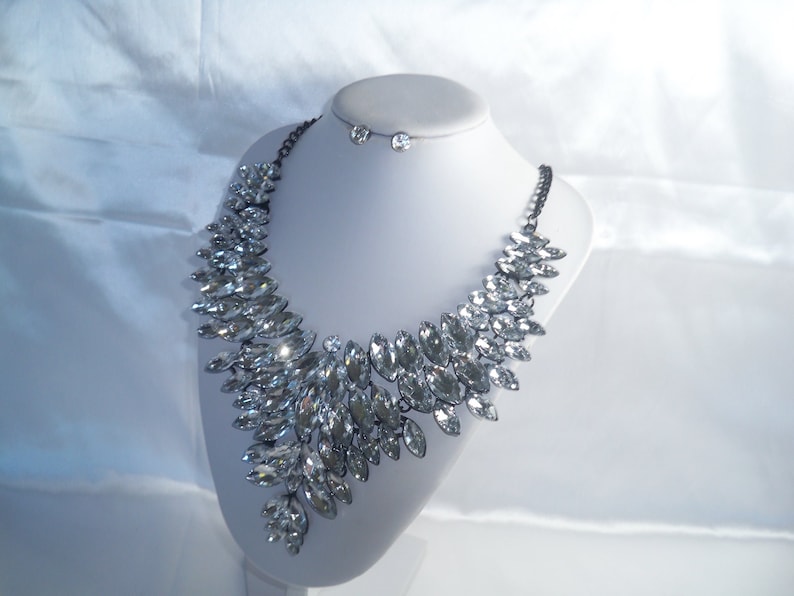 Puede incluir: Un collar llamativo con numerosos cristales transparentes de talla marquesa dispuestos en un patr&oacute;n denso y superpuesto. Los cristales est&aacute;n engastados en un marco de metal oscuro con una cadena a juego. Se incluye un par de pendientes de cristal redondos.