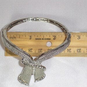 May include: Silver-toned bracelet with a bow design. The bracelet is adorned with small, sparkling rhinestones. The bow has a pearl-like center. The bracelet is approximately 2 inches in diameter.