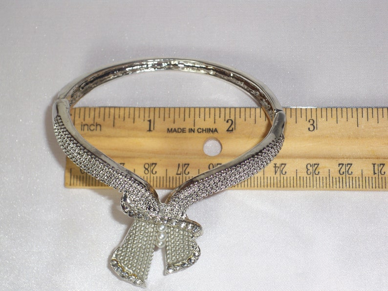 May include: Silver-toned bracelet with a bow design. The bracelet is adorned with small, sparkling rhinestones. The bow has a pearl-like center. The bracelet is approximately 2 inches in diameter.