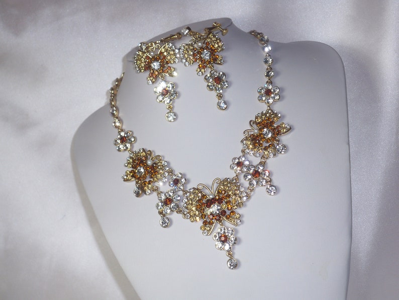May include: A gold necklace and earrings set with clear and amber rhinestones. The necklace features a butterfly design with multiple crystal accents.