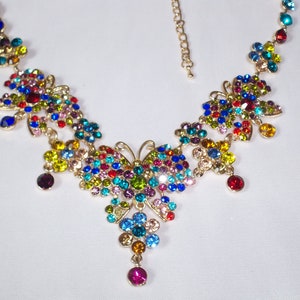 Puede incluir: Un collar de mariposa de colores con una cadena dorada y piedras de cristal multicolores. El collar presenta un dise&ntilde;o de mariposa grande con peque&ntilde;os detalles de mariposa.