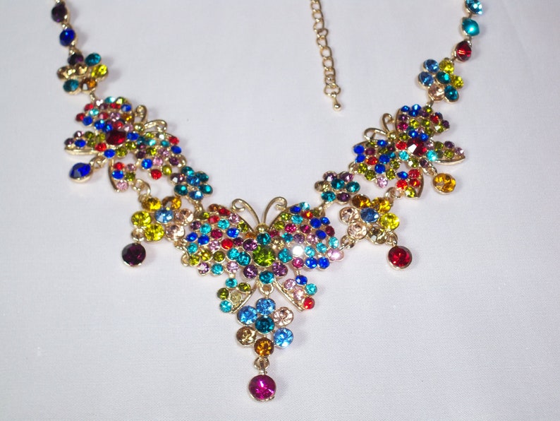 Puede incluir: Un collar de mariposa de colores con una cadena dorada y piedras de cristal multicolores. El collar presenta un dise&ntilde;o de mariposa grande con peque&ntilde;os detalles de mariposa.