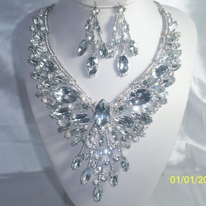 May include: A silver crystal necklace and earrings set. The necklace features a large, teardrop-shaped crystal pendant surrounded by smaller crystals. The earrings are also teardrop-shaped and dangle from silver hooks.