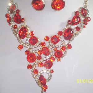 May include: A gold-coloured necklace and matching earrings set. The necklace has a cascading design of large, oval, and round red gemstones, with smaller clear crystals. The earrings are round with a large red gemstone.