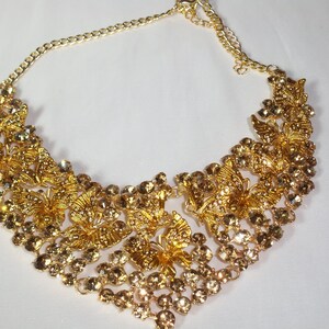 Puede incluir: Un collar dorado con intrincados dise&ntilde;os de mariposas y brillantes pedrer&iacute;a transparente y &aacute;mbar. El collar tiene una delicada cadena y un colgante tipo babero, perfecto para ocasiones especiales.