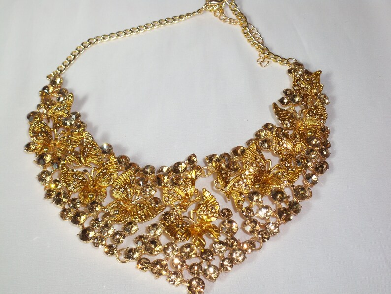 Puede incluir: Un collar dorado con intrincados dise&ntilde;os de mariposas y brillantes pedrer&iacute;a transparente y &aacute;mbar. El collar tiene una delicada cadena y un colgante tipo babero, perfecto para ocasiones especiales.
