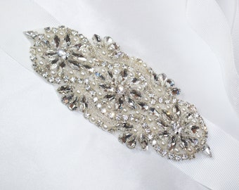 May include: A white satin ribbon with a crystal and pearl embellishment. The embellishment features a floral design with clear crystals and white pearls.