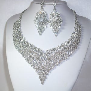 May include: A silver-toned necklace and earring set. The necklace features a cascading design of clear, sparkling crystals. The matching earrings are a similar design. The jewelry is displayed on a white mannequin bust.