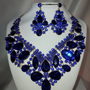 May include: A striking blue jewellery set, comprising a necklace and matching earrings, presented on a white mannequin. The necklace and earrings feature large, faceted blue gemstones set in a gold-toned metal. The earrings are a cluster design.