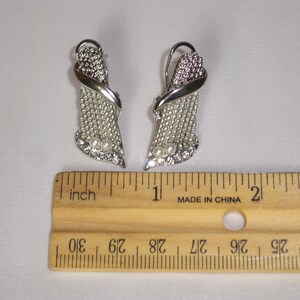 May include: A pair of silver-toned earrings with a decorative design. Each earring features a textured, openwork section, accented with clear rhinestones and two small, round pearls. The earrings are approximately 1 inch in length.
