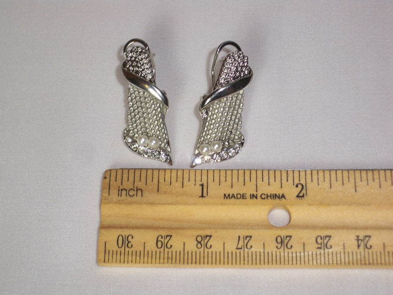 May include: A pair of silver-toned earrings with a decorative design. Each earring features a textured, openwork section, accented with clear rhinestones and two small, round pearls. The earrings are approximately 1 inch in length.